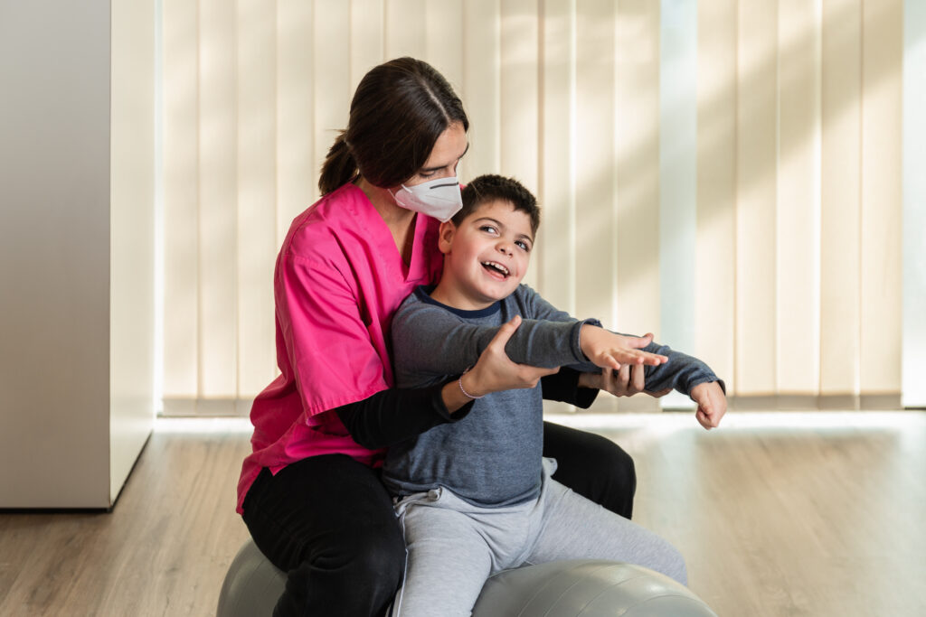 child with multiple intellectual and physical disabilities and physiotherapist woman do balance exercises on a Peanut Gym Ball with joy.