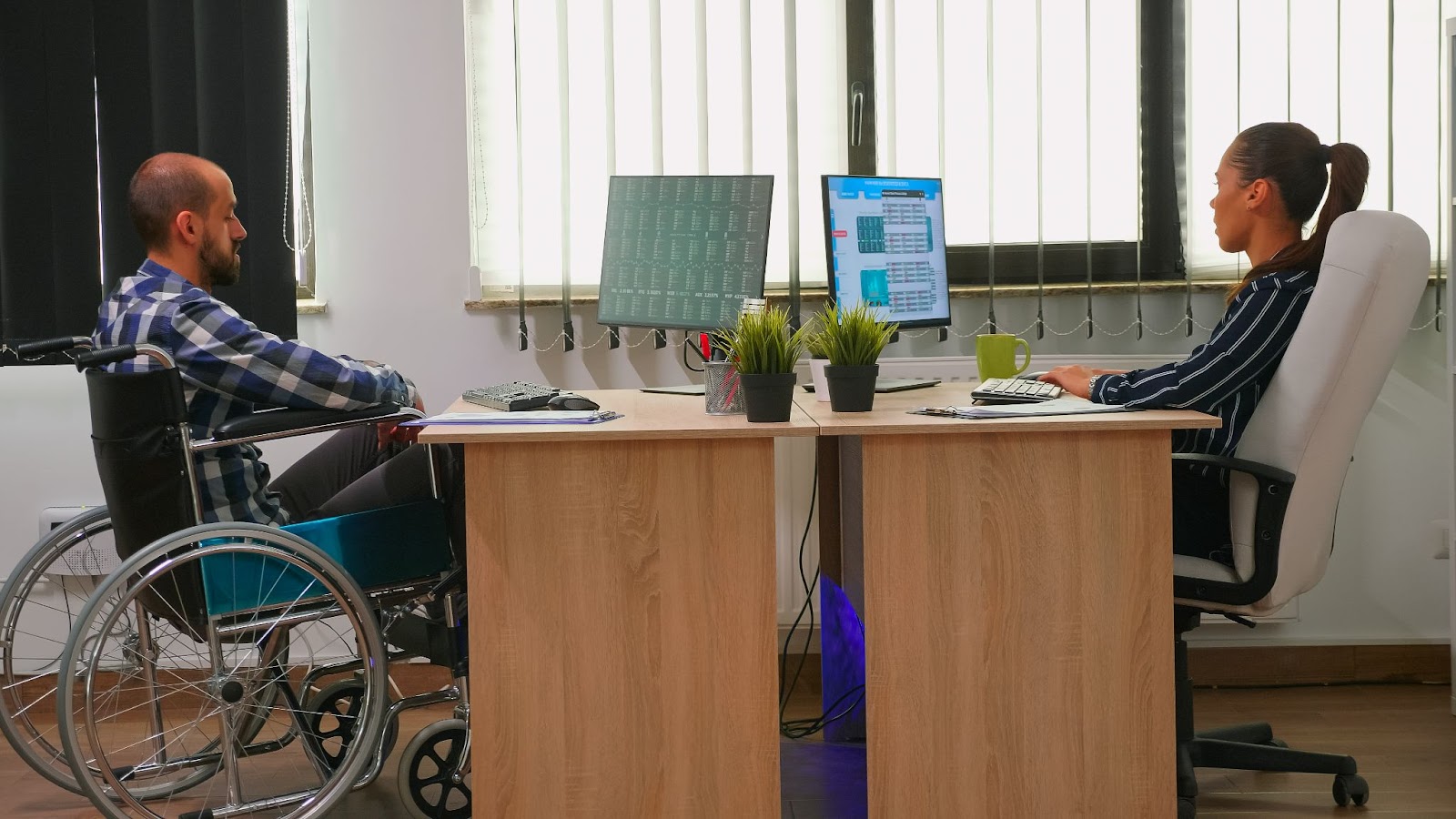 Man in a wheelchair working at a desk with a colleague, showcasing an accessible home modifications setting